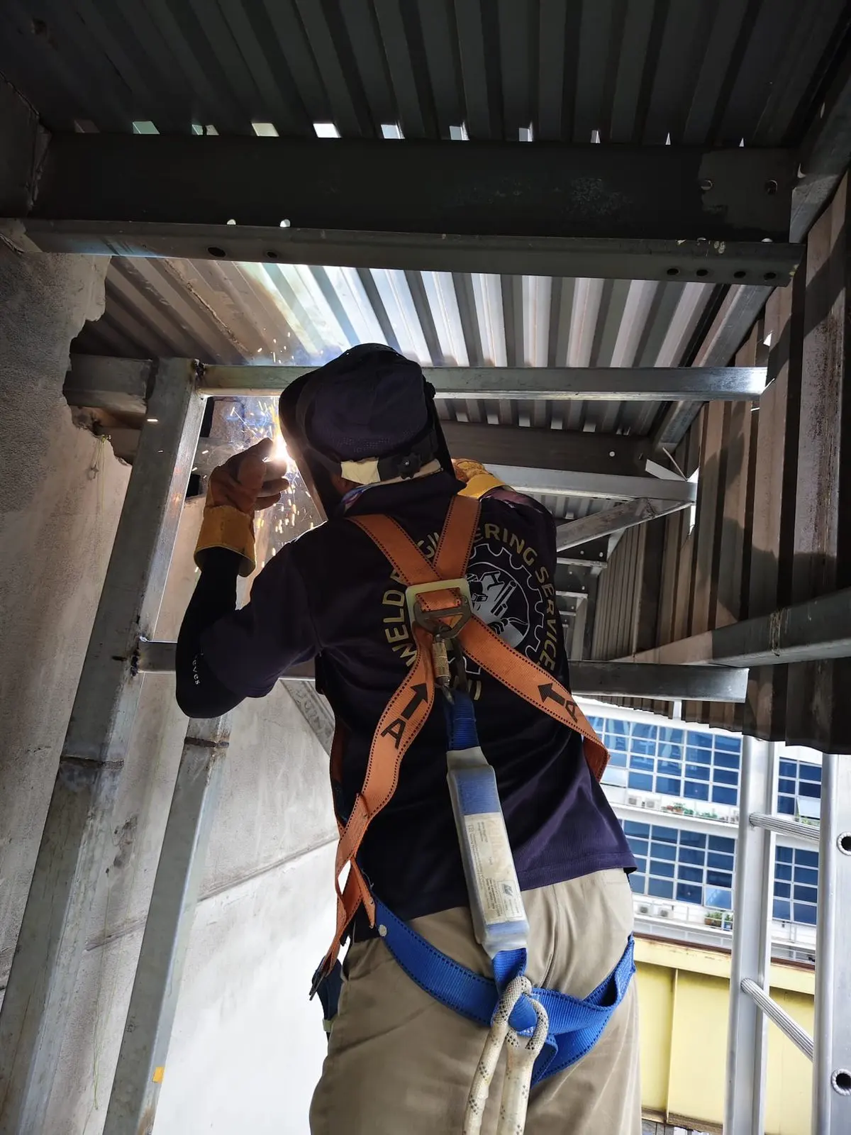 Fabricator welding the cat ladder structure on site at Buroh Street while wearing a full-body harness with lanyard for fall protection